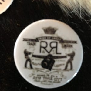 K&ouml;nnte beinhalten: Ein wei&szlig;er Knopf mit einem schwarz-wei&szlig;en Design. Das Design zeigt eine Krone, die Buchstaben "R&L", eine Kamera und den Text "Trade Mark" und "New York City".