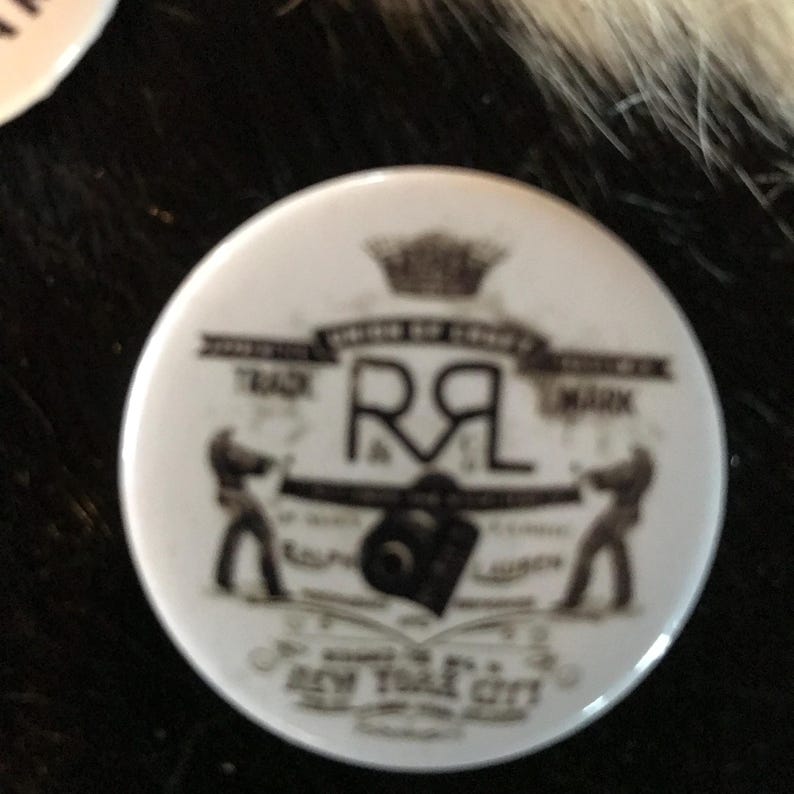 K&ouml;nnte beinhalten: Ein wei&szlig;er Knopf mit einem schwarz-wei&szlig;en Design. Das Design zeigt eine Krone, die Buchstaben "R&L", eine Kamera und den Text "Trade Mark" und "New York City".