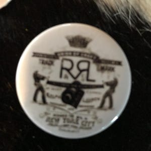 K&ouml;nnte beinhalten: Ein wei&szlig;er Knopf mit einem schwarz-wei&szlig;en Design. Das Design zeigt eine Krone, die Buchstaben "RL", eine Kamera und den Text "New York City".