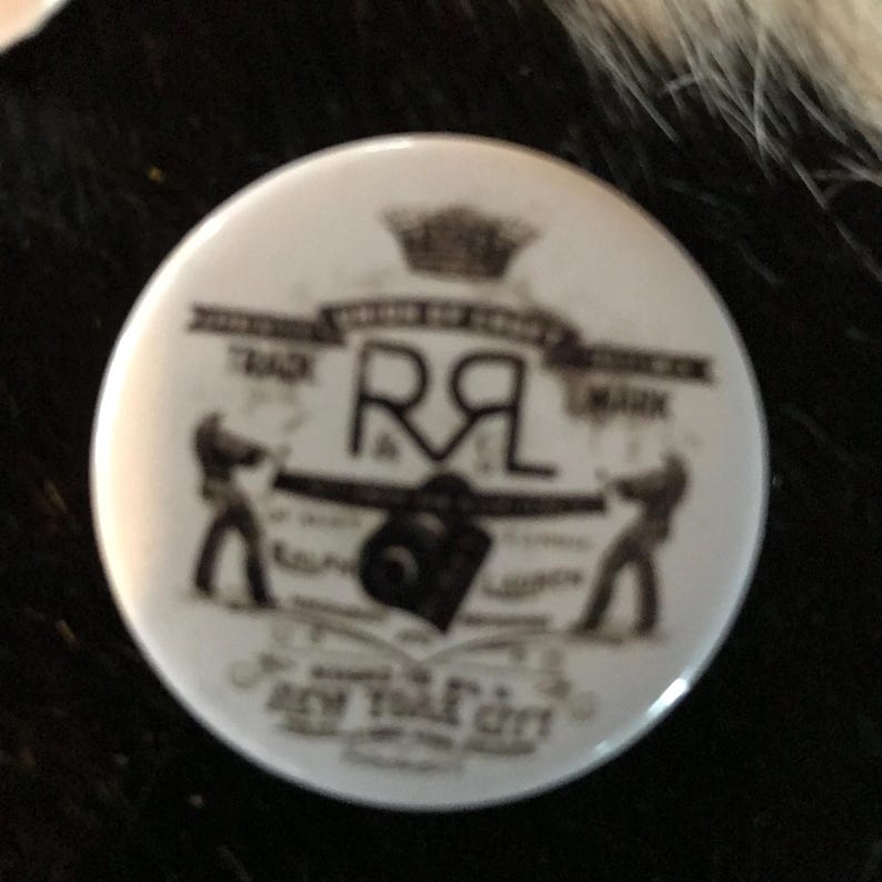 K&ouml;nnte beinhalten: Ein wei&szlig;er Knopf mit einem schwarz-wei&szlig;en Design. Das Design zeigt eine Krone, die Buchstaben "RL", eine Kamera und den Text "New York City".