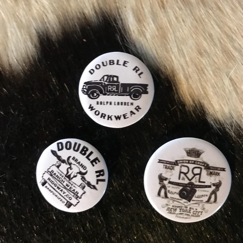 K&ouml;nnte beinhalten: Drei wei&szlig;e runde Kn&ouml;pfe mit schwarzem Text und Bildern. Der obere Knopf sagt "Double RL Ralph Lauren Workwear" und zeigt ein schwarzwei&szlig;es Bild eines Pick-up-Trucks. Der linke Knopf sagt "Double RL Brand" und zeigt ein schwarzwei&szlig;es Bild eines Bullenkopfes und ein Schild mit der Aufschrift "Genuine Ranch Wear Ridgway.Co Sanforized". Der rechte Knopf sagt "Trade Mark" und zeigt ein schwarzwei&szlig;es Bild von zwei M&auml;nnern, die eine Kamera halten, mit dem Text "Ralph Lauren New York City".