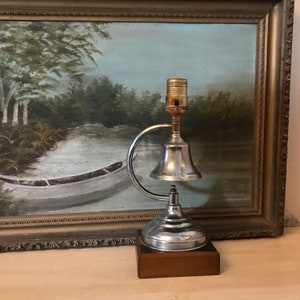 May include: A vintage silver bell lamp with a wooden base. The lamp has a curved arm and a single light bulb. The lamp is in front of a framed painting of a river scene.