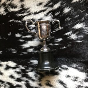 May include: A silver trophy cup with two handles and a black base. The cup has engraved text that reads "UNDERSKATE 1938, W.J. LESLIE, 2ND, J.A. MAINE CUP". The trophy is set against a black and white patterned background.