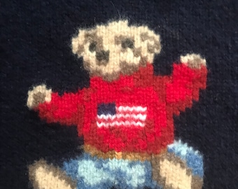 May include: Navy blue sweater featuring a pixelated teddy bear wearing a red sweater with an American flag design and blue jeans. The brand name "RALPH LAUREN" is visible at the top and bottom of the sweater.
