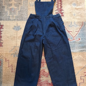 May include: Dark blue denim overalls with wide legs and a bib top. The overalls have adjustable straps, pleats at the waist, and button details on the sides. The garment is made of a sturdy denim fabric.