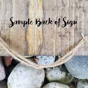 May include: The back of a wooden sign with the text "Sample Back of Sign" written in black cursive. The sign has two metal hooks with twine attached to each hook.
