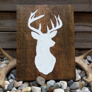 May include: A rustic wooden sign featuring a white silhouette of a deer head with antlers. The sign is rectangular and has a dark brown wood grain. Antlers and stones are in the foreground.
