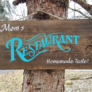 May include: A brown wooden sign with the text "Mom's Restaurant" in turquoise blue lettering and "Homemade Taste!" in white lettering.