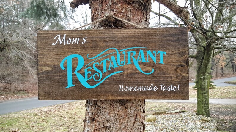 May include: A brown wooden sign with the text "Mom's Restaurant" in turquoise blue lettering and "Homemade Taste!" in white lettering.