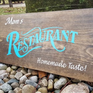 May include: A wooden sign with the text "Mom's Restaurant Homemade Taste!" in white lettering. The word "Restaurant" is in a stylized blue font.