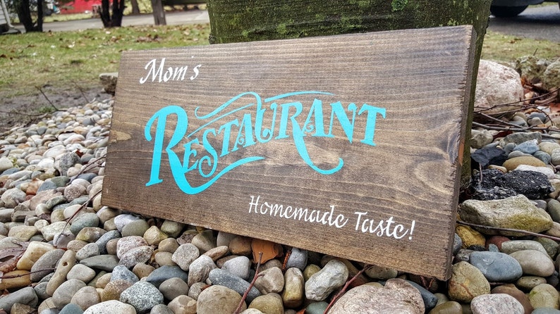 May include: A wooden sign with the text "Mom's Restaurant Homemade Taste!" in white lettering. The word "Restaurant" is in a stylized blue font.