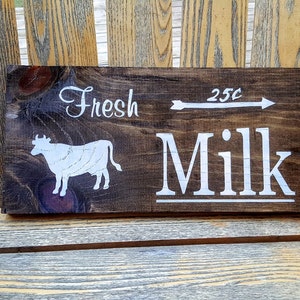 May include: A rustic wooden sign with white painted text that reads "Fresh 25c Milk" with a white silhouette of a cow. The sign is dark brown with a distressed finish.