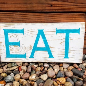 May include: A white wooden sign with the word "EAT" painted in turquoise blue letters. The sign is sitting on a bed of small, colorful rocks.