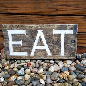 May include: A rustic wooden sign with the word "EAT" painted in white on a dark brown background. The sign is sitting on a bed of small, colorful rocks.