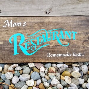 May include: A brown wooden sign with the text "Mom's Restaurant" in turquoise lettering and "Homemade Taste!" in white lettering.