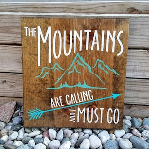 May include: A wooden sign with the text "The Mountains Are Calling And I Must Go" in white paint. The sign also features a turquoise mountain silhouette and a turquoise arrow pointing to the right.