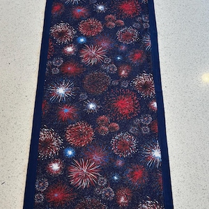 May include: A rectangular table runner with a navy blue border and a pattern of red, white, and blue fireworks on a dark blue background. The runner is approximately 12 inches wide and 36 inches long.