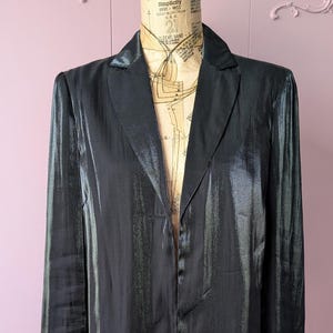 May include: A long, black, shiny coat with a notched collar and long sleeves. The coat is displayed on a mannequin with a vintage sewing pattern. The coat has a single-button closure and a sleek, flowing design, suitable for formal occasions.