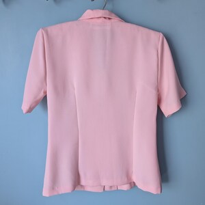 May include: A pink short-sleeved collared blouse with a button-up front. The blouse has a fitted silhouette and a slightly flared hem.