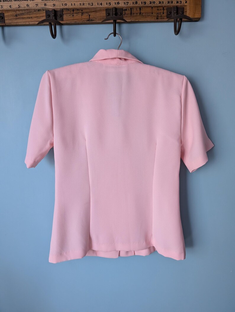 May include: A pink short-sleeved collared blouse with a button-up front. The blouse has a fitted silhouette and a slightly flared hem.
