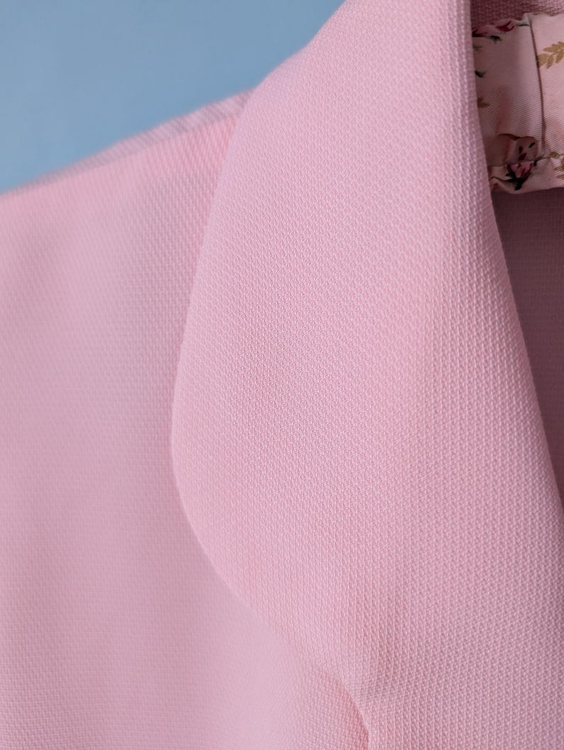 May include: A close-up of a pink blazer with a textured fabric. The blazer has a notched collar and a single button closure.