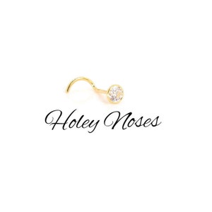 Puede incluir: Un piercing de nariz dorado con un poste curvo y una gema redonda y transparente. El texto "Holey Noses" est&aacute; escrito en cursiva debajo del piercing sobre un fondo blanco.