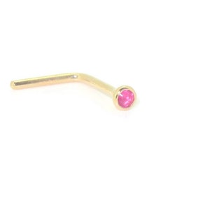 May include: A gold nose stud featuring a small, round, pink gemstone. The L-shaped stud has a straight post and a bezel-set gemstone. The jewellery is set against a white background.