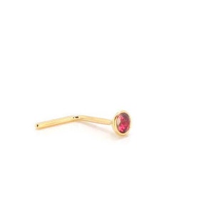 Może przedstawiać: Złoty kolczyk do nosa z małym, okrągłym, czerwonym kamieniem szlachetnym. Kolczyk ma prosty pręt, który zgina się pod kątem prostym, z kamieniem osadzonym w oprawie. Biżuteria jest umieszczona na białym tle.