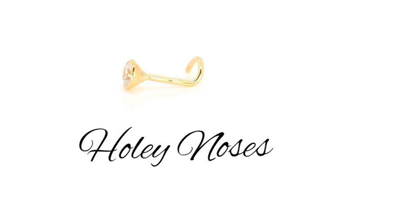 Puede incluir: Un piercing de nariz dorado con una gema clara y redonda. El piercing es curvo y tiene un peque&ntilde;o engaste decorativo. El texto "Holey Noses" est&aacute; escrito en cursiva debajo.
