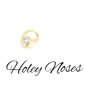 Puede incluir: Un piercing de nariz dorado con una gema redonda y transparente engastada en un bisel. El piercing tiene un poste curvo. El texto "Holey Noses" est&aacute; escrito en cursiva debajo.
