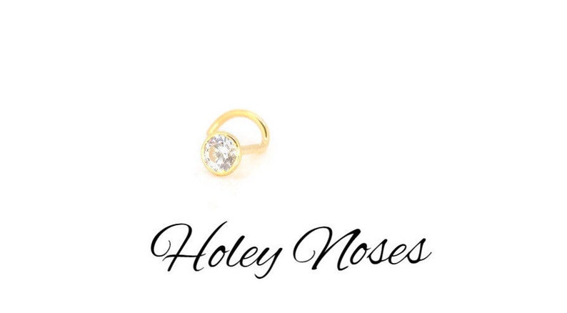 Puede incluir: Un piercing de nariz dorado con una gema redonda y transparente engastada en un bisel. El piercing tiene un poste curvo. El texto "Holey Noses" est&aacute; escrito en cursiva debajo.