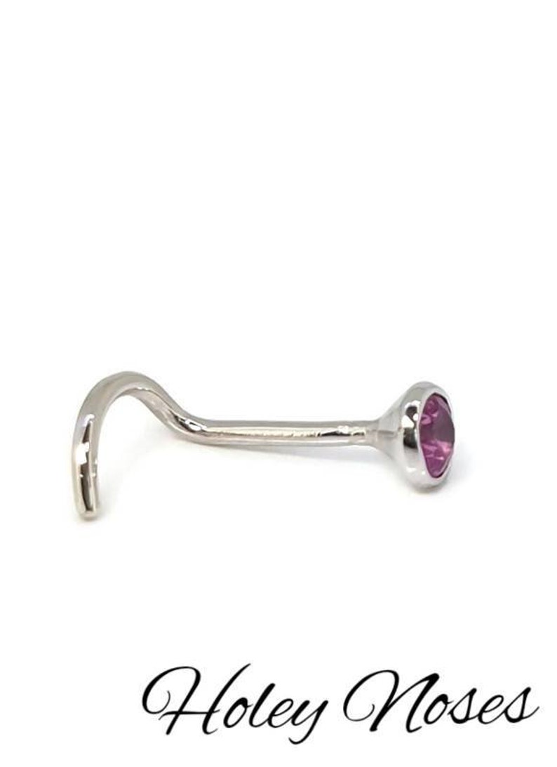 May include: A silver nose stud with a curved post and a bezel-set, round, pink gemstone. The stud is designed with a curved end for secure placement. The text "Holey Noses" is at the bottom.