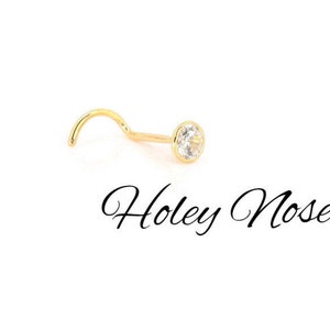 Puede incluir: Un piercing de nariz dorado con una gema clara y redonda. El piercing tiene un poste curvado y un engaste biselado. El texto "Holey Noses" est&aacute; escrito en cursiva debajo.