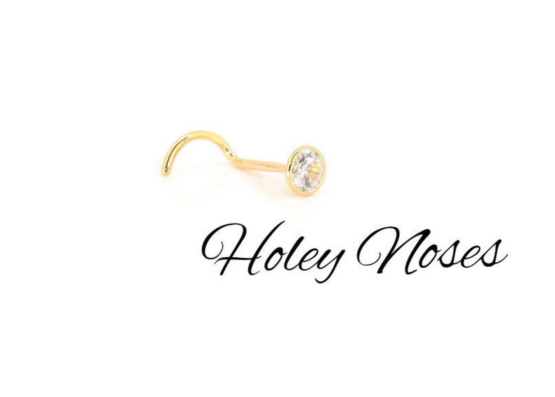 Puede incluir: Un piercing de nariz dorado con una gema clara y redonda. El piercing tiene un poste curvado y un engaste biselado. El texto "Holey Noses" est&aacute; escrito en cursiva debajo.