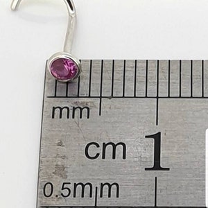 May include: A silver nose ring with a curved post and a round, pink gemstone. The ring is shown next to a ruler, with the gemstone appearing to be approximately 0.2 cm in diameter.