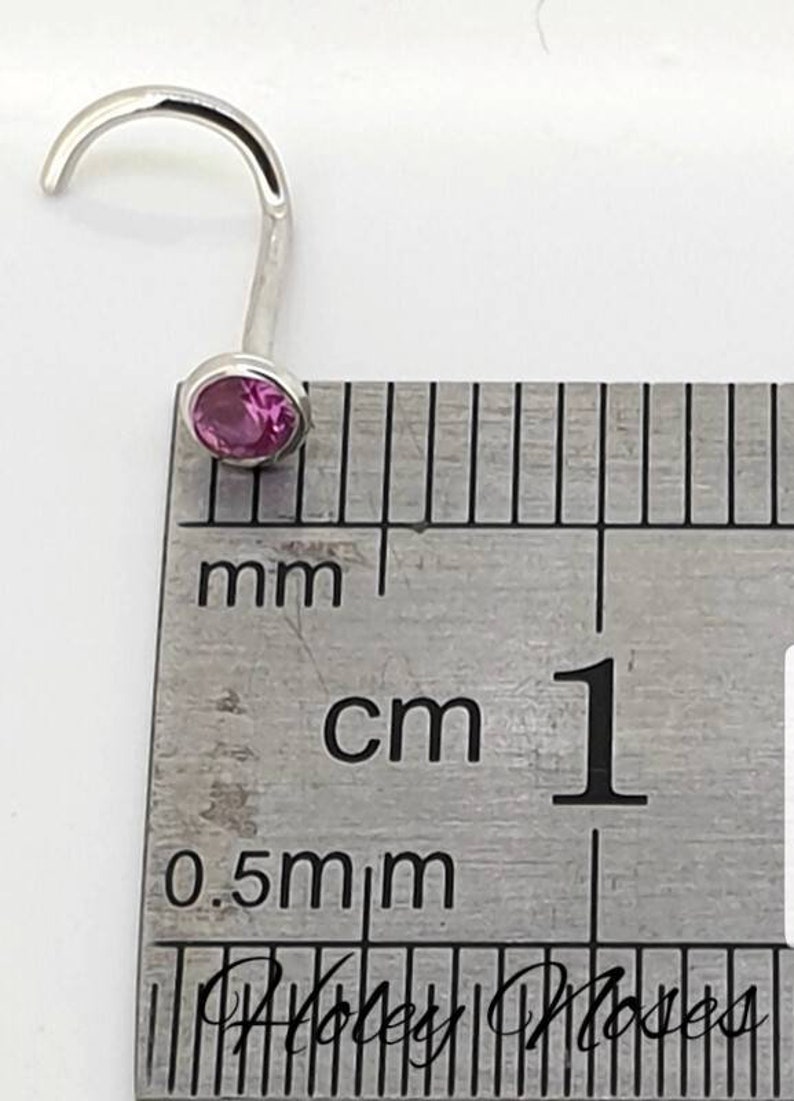 May include: A silver nose ring with a curved post and a round, pink gemstone. The ring is shown next to a ruler, with the gemstone appearing to be approximately 0.2 cm in diameter.