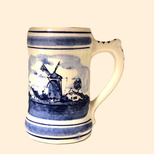 Könnte beinhalten: Ein Bierkrug aus Keramik mit blau-weißem Windmühlen-Design. Der Krug hat einen Henkel und ist mit einer Landschaftsszene einer Windmühle, Häusern und Wasser verziert. Ober- und Unterseite des Kruges sind mit blauen Streifen versehen.