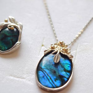 Könnte beinhalten: Ein silbernes Halsketten- und Ohrring-Set. Der Anhänger und die Ohrringe zeigen schillernde blau-grüne ovale Steine, die in Silber mit goldfarbenen Akzenten gefasst sind. Der Schmuck wird in einer weißen Schachtel präsentiert.