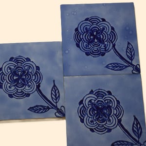 Può includere: Quattro piastrelle quadrate blu con un motivo floreale. Ogni piastrella presenta un fiore blu scuro con uno stelo e foglie. Le piastrelle sono disposte per mostrare il motivo. Lo sfondo è di colore beige chiaro.