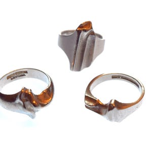 Könnte beinhalten: Drei silberfarbene Metallringe mit unregelmäßigen, skulpturalen Oberflächen. Jeder Ring hat eine einzigartige, organische Form mit glatten, reflektierenden Bereichen und vertieften Details. Im Inneren eines Rings ist das Wort LAPPONIA eingraviert.