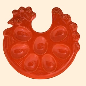 May include: A red, hen-shaped egg holder. The plastic tray has six egg-shaped indentations and a handle. The hen's head and tail are detailed with textured ridges. The tray is a solid, glossy red color.