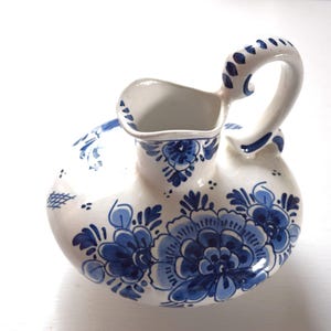 Może przedstawiać: Biało-niebieski ceramiczny dzbanek z zakrzywionym uchwytem i szerokim wylewem. Dzbanek jest ozdobiony niebieskimi wzorami kwiatowymi. Uchwyt i wylew również zdobione są niebieskimi akcentami. Prawdopodobnie służy do serwowania płynów.