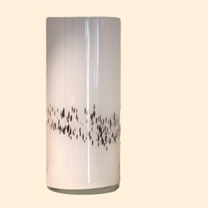 Könnte beinhalten: Eine zylindrische Vase aus weißem Glas mit einem Muster aus schwarzen Strichen in der Mitte. Die Vase ist hoch und schlank, geeignet für Blumen oder als dekoratives Element. Der Hintergrund ist hellbeige. Etwa 20 cm hoch.