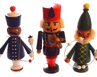 Könnte beinhalten: Drei Holz-Nussknacker in Form von Figuren. Die Nussknacker sind in verschiedenen Farben bemalt, darunter Marineblau, Rot und Grün. Eine Figur hat einen roten Hut und raucht eine Pfeife. Die anderen beiden Figuren halten Zubehör.