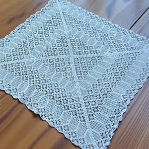 May include: A square, off-white crocheted doily with a detailed floral and geometric pattern. The edges are scalloped, and the design features repeating diamond and flower motifs. The doily is placed on a wooden surface.