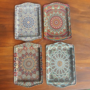 May include: Four rectangular decorative trays with ornate, colorful mandala designs. The trays feature a variety of colors, including blue, red, green, and gold, and are arranged on a wooden surface. Each tray has a unique pattern.