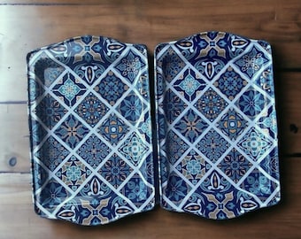 May include: Two rectangular serving trays with a decorative, repeating pattern. The pattern features blue, teal, and gold geometric shapes and floral motifs on a white grid. The trays have a curved top edge and are set on a wooden surface.