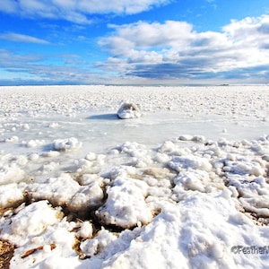 May include: A scenic landscape of a frozen lake under a bright blue sky with scattered clouds. The foreground features large chunks of ice and snow, transitioning to a vast expanse of white ice in the distance.