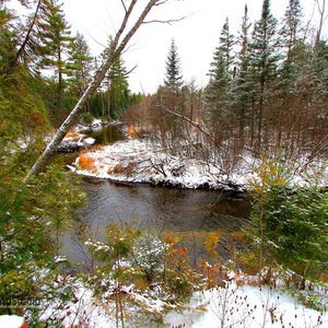 May include: A scenic landscape photograph of a river winding through a snow-covered forest. Evergreen trees and bare branches frame the water, with patches of snow on the banks and ground. The sky is overcast.