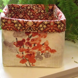 May include: A fabric storage basket with a floral design. The exterior is cream-coloured with orange and grey floral patterns. The top and interior lining have a patterned design in shades of brown, orange, and cream.